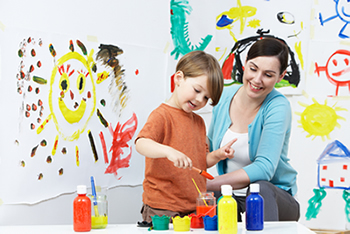 A female adult smiles watching a young boy paint his mural on paper