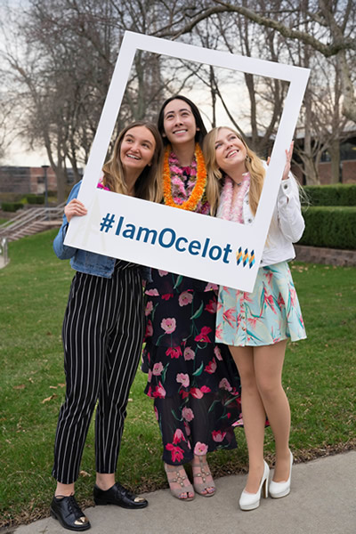 Three women, the Pythagorean Prize winners, pose in a poster frame that reads hashtag I am ocelot