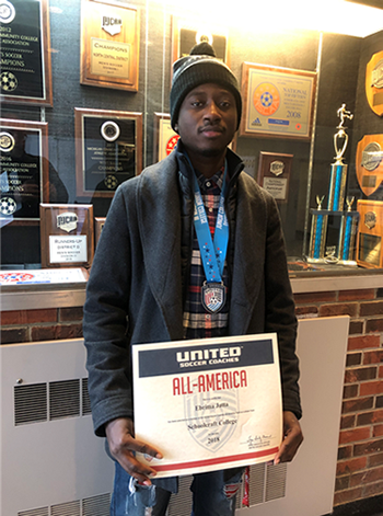 Soccer player Ebrima Jatta holds a certificate in front of a trophy showcase that reads "All-American" title