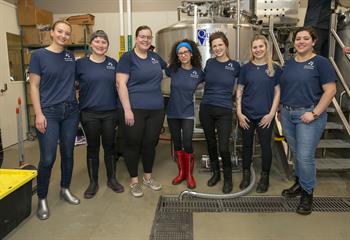 A group of young women pose for a photo within a brewing distillation area.