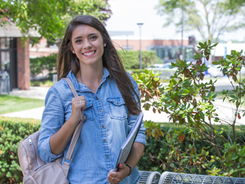 A young female student smiling and holding class books, standing outside with greenery campus backdrop